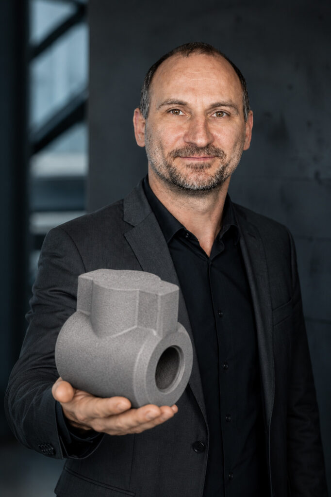 Ein Mann in einem dunklen Anzug steht in einem Haus, lächelt leicht und hält ein großes, graues, industriell aussehendes 3D-gedrucktes Objekt in die Kamera. Der Hintergrund ist unscharf und modern.