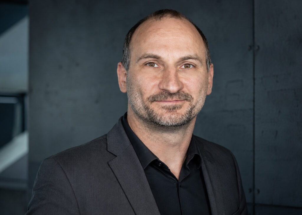 Ein Mann mit kurzen braunen Haaren, einem kurzgeschnittenen Bart und Schnurrbart, der eine dunkle Anzugsjacke und ein schwarzes Hemd trägt, steht vor einem dunklen, strukturierten Hintergrund und lächelt leicht in die Kamera.